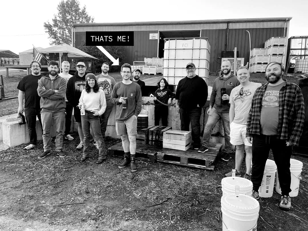 A homebrew club, in black and white, standing in front of a cider press barn, with fermenter buckets and totes around them. An arrow labeled “That’s me” points at Chris, in the middle, holding a beer.